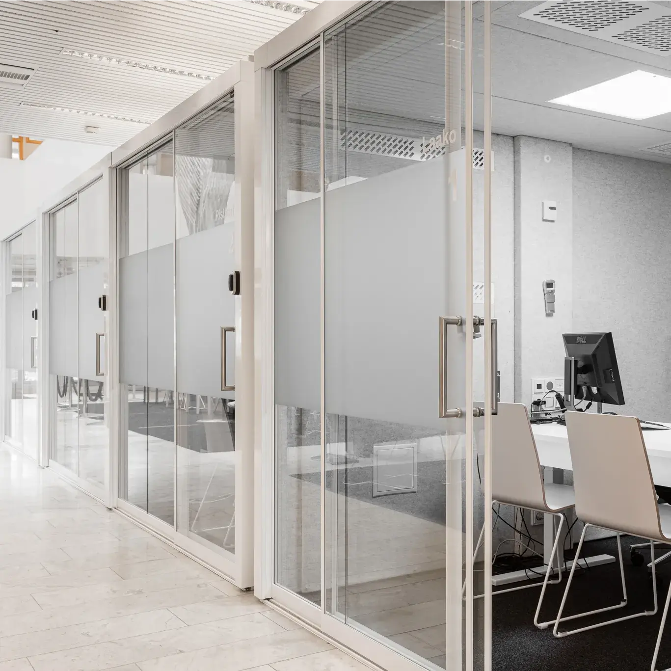 Bright office corridor with glass-walled meeting pods featuring frosted panels, sliding doors, and minimalist furniture inside.