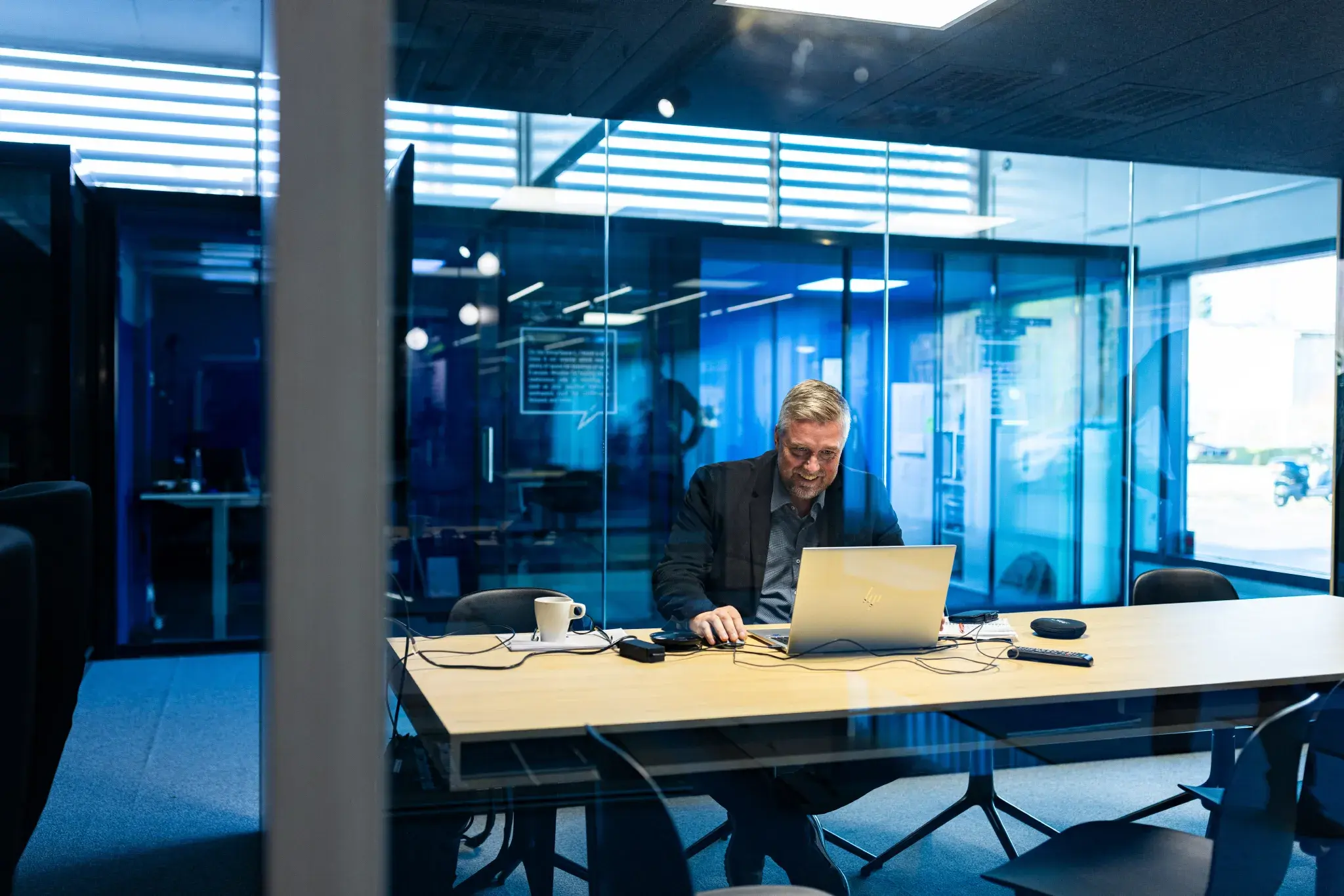 Man working on a laptop at a conference table inside a glass-walled office pod, with modern office furnishings and blue-toned lighting.